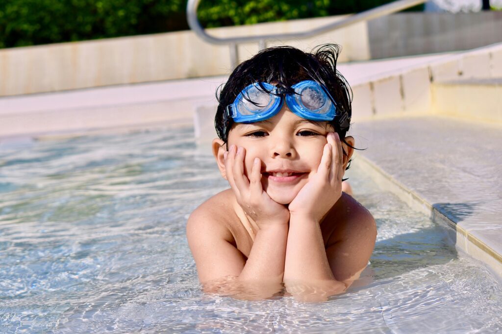 Foto de uma criança sorrindo na piscina com óculos de natação, simbolizando os benefícios da natação infantil para a saúde.